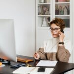 Smart confident female managing director listening to employees report on smartphone while working on computer in light modern office