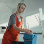 A woman in a gray shirt and red overalls cleans a kitchen counter using a blue bucket and white rag. Modern setting.